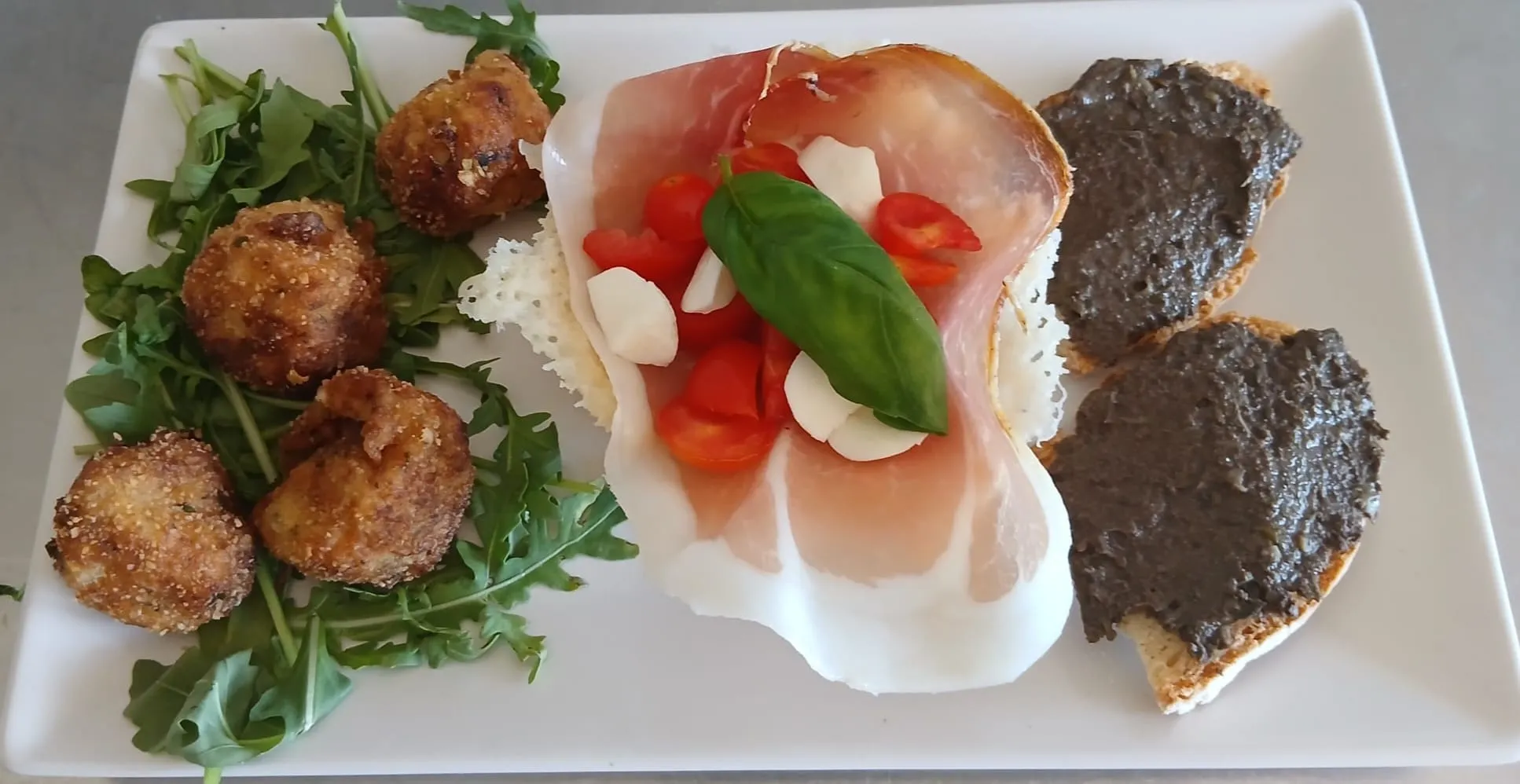 Antipasto misto: polpette fritte su un letto di rucola, prosciutto crudo con pomodorini e mozzarella, e crostini al patè.
